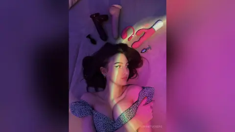Young woman in a floral patterned dress lying on a bed under moody pink and blue neon lighting.