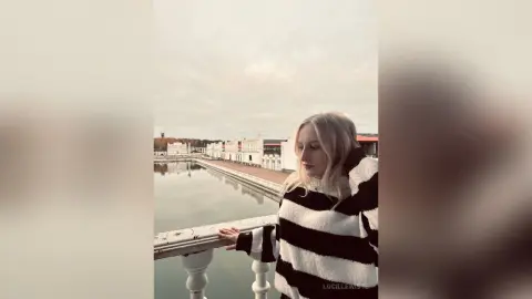 Blonde woman in a black and white striped sweater standing by a scenic lake under a cloudy sky.