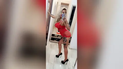 Tattooed woman in a red lace bodysuit and black high heels taking a mirror selfie in a bright modern room.