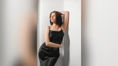 Elegant woman in a black satin slip dress leaning against a white wall in a minimalist, moody setting.