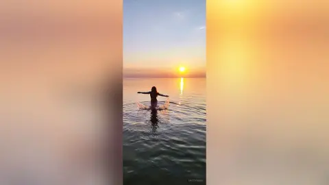 Silhouette of a woman in a bikini standing in calm ocean water during a peaceful golden sunset.