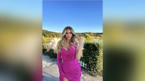 TiffanyAddams poses outdoors in a bright pink, low-cut ruched dress with long sleeves, letting her blonde hair fall over her shoulders.