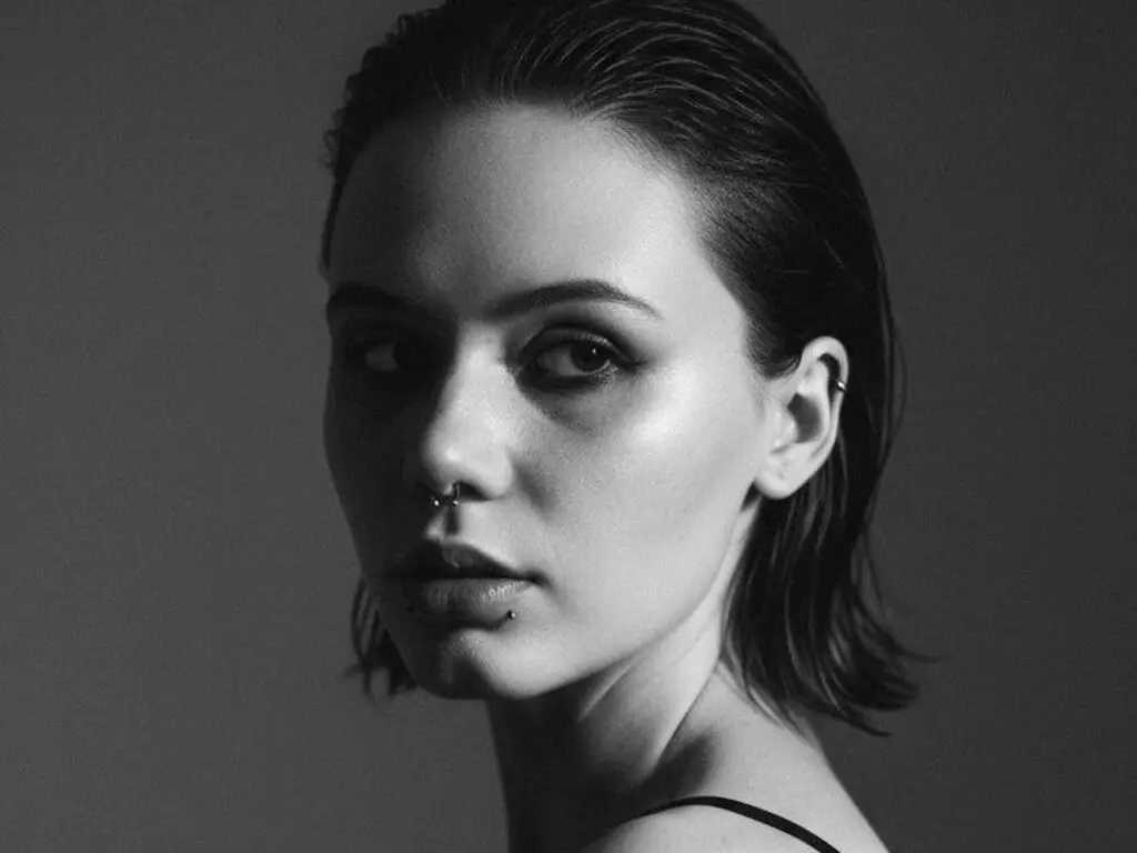 Moody black and white portrait of a young woman with slicked-back hair and a nose piercing in soft studio lighting.