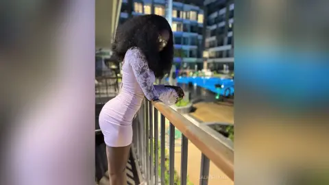 Woman in a tight pink mini dress leaning on a balcony railing at night in a modern urban setting.