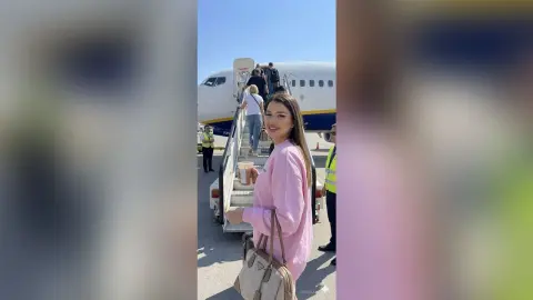 CapriAndersen smiles while standing outside on a sunny day, wearing a pink long-sleeve sweatshirt and holding a drink and a tan handbag near an airplane.