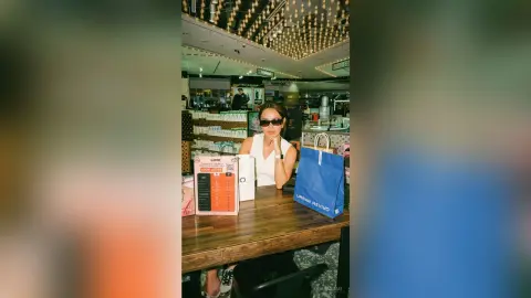 Stylish woman in a white sleeveless top sitting at a cafe table with blue and white shopping bags.
