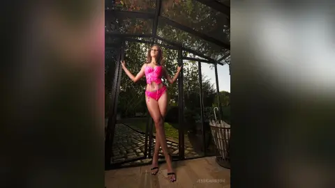 Confident woman in a pink bikini posing in a sunlit glass greenhouse garden.