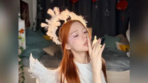 Redheaded woman in white angel costume and feathered wings posing in a cozy bedroom setting.