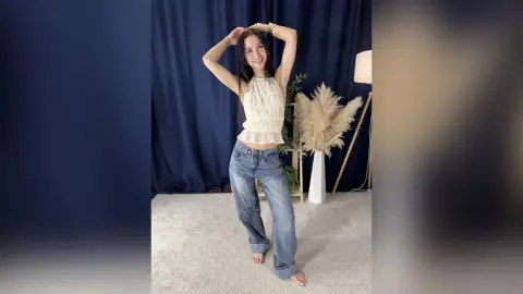 Smiling young woman in a white ruffled halter top and blue jeans posing in a bright studio setting.