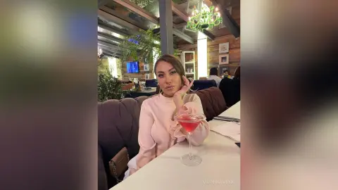 VictoriaWynn poses at a restaurant table wearing a light pink long-sleeved top, holding a pink cocktail glass near her face.