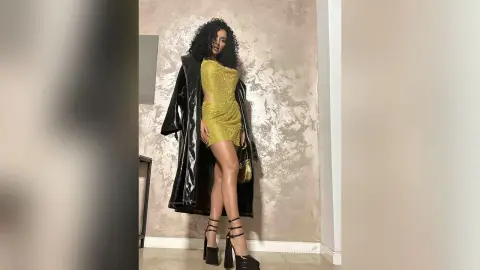NicoleBella stands in a gold glittery mini dress and black strappy high heels against a textured wall.