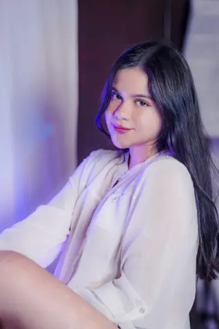 Young woman in a white button-down shirt smiling softly in a dreamy, blue-lit indoor setting.