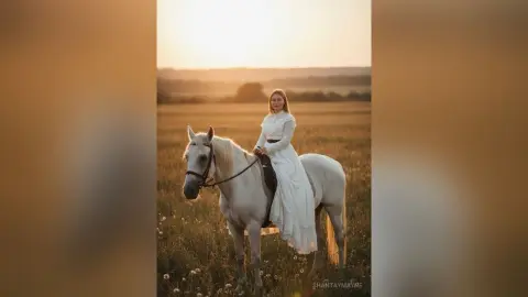 ShantayMayne sits upright on a white horse in a golden field wearing a long white dress.