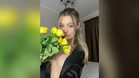Blonde woman in a black satin outfit holding a bouquet of yellow roses in a cozy indoor setting.