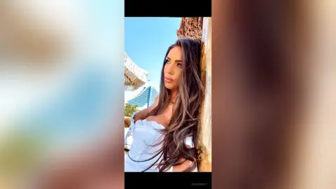 ArianaKroft poses outdoors with her long brown hair flowing, wearing a white off-the-shoulder top against a bright blue sky.