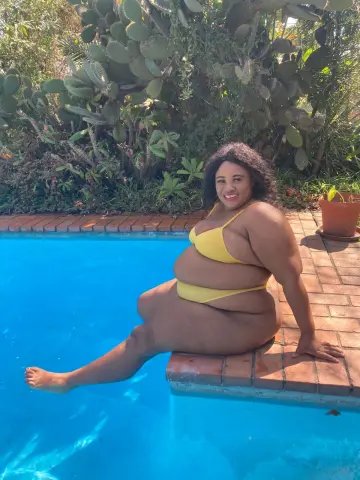 Smiling curvy woman in a yellow bikini relaxing by a bright blue swimming pool in a lush garden setting.