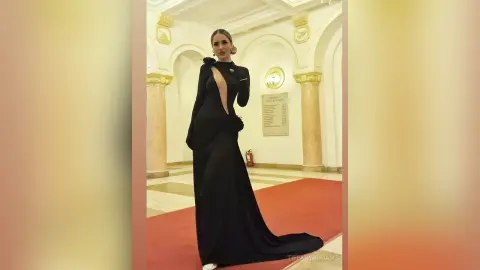 TiffanyAddams poses in a long black dress with a deep cutout, standing in a grand hall with white arches and a red carpet.