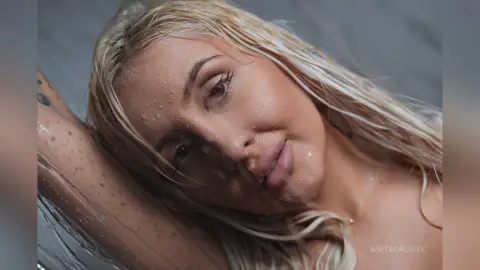 Anita poses with wet blonde hair and glistening skin against a grey background.
