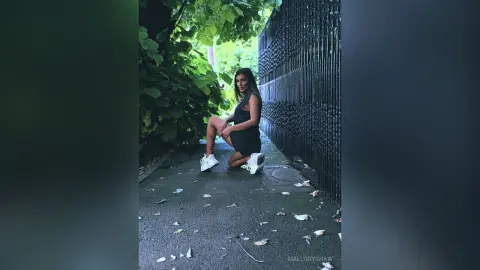 Stylish woman in a black romper and white sneakers posing on a leafy urban path next to a dark textured wall.
