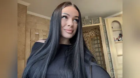 AriaVex looks off to the side with her long, straight black hair falling over her shoulders while wearing a black top.