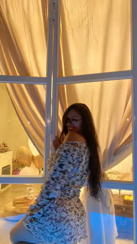 Smiling woman in a floral off-the-shoulder dress posing by a window with warm indoor lighting.