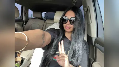 Woman with long dark hair and sunglasses wearing a black t-shirt sitting in a car interior.