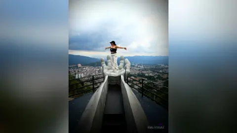 Woman in black crop top standing on giant stone hands overlooking a scenic mountain valley under a cloudy sky.