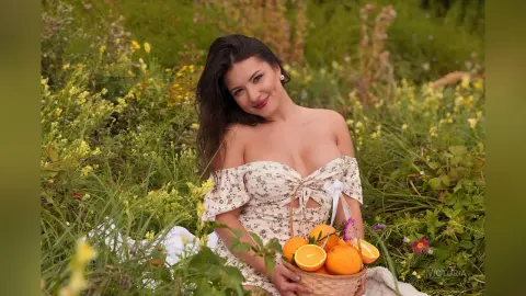 Smiling woman in a floral off-the-shoulder dress sitting in a sunny wildflower meadow with a basket of oranges.