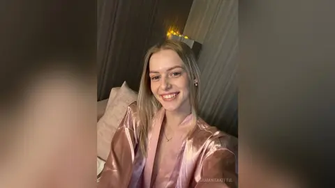Smiling blonde woman in a pink satin robe relaxing in a cozy, dimly lit bedroom.