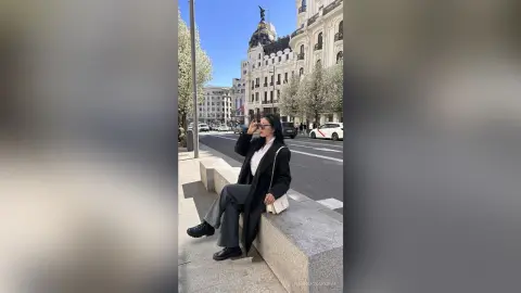 NashiroSakurai sits on a stone ledge wearing a black coat, white shirt, and dark trousers while wearing sunglasses outdoors.
