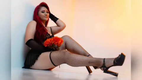 Red-haired woman in black lace lingerie and fishnet tights sitting against a wall in warm studio lighting.