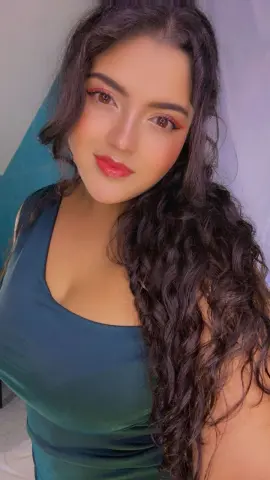 Young woman with long wavy hair wearing a dark teal satin dress in a soft indoor setting.