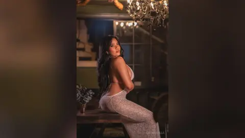 DayanaHille poses sideways in a white lace bodysuit, showing her back and long dark hair against a dark background.