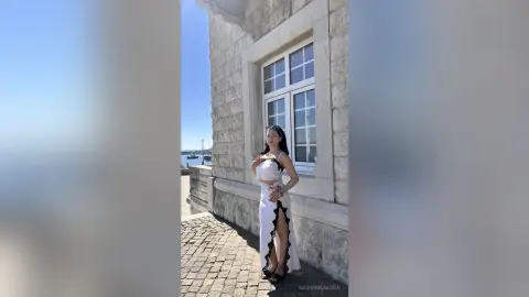Elegant woman in a white cutout maxi skirt and black crop top standing by a stone building on a sunny day.