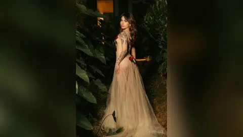 Elegant woman in a flowing beige tulle gown posing in a lush, moody tropical garden at twilight.