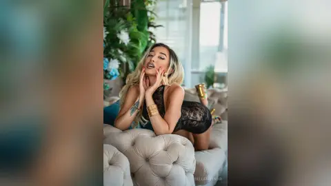 Glamorous blonde woman in a black lace bodysuit relaxing on a beige sofa in a bright, modern living room.