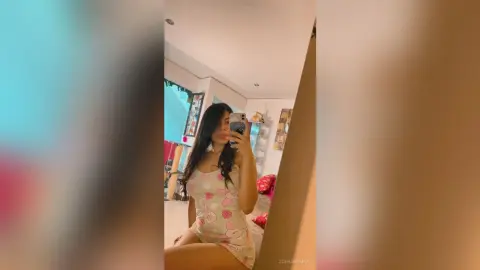 Young woman in a white floral mini dress taking a mirror selfie in a bright bedroom setting.