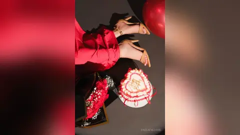 Woman in red silk outfit sitting by a heart-shaped cake and roses for a romantic celebration.