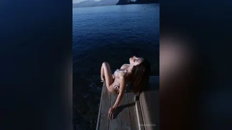 Relaxed woman in a white bikini lounging on a wooden pier by the deep blue ocean at twilight.