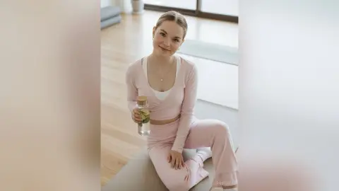 Smiling young woman in pink loungewear sitting in a bright modern living room holding a glass water bottle.