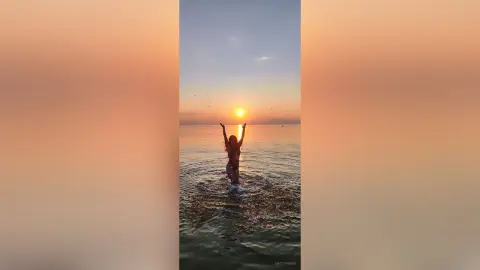 Silhouette of a woman in a bikini raising arms during a peaceful golden sunset over the ocean waves.