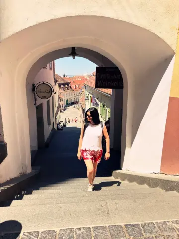 Woman in a white tank top and red shorts walking down stone stairs under a sunny European archway.