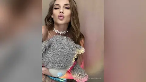 Glamorous woman in a sparkling necklace holding a large grey plush teddy bear in a soft-lit indoor setting.