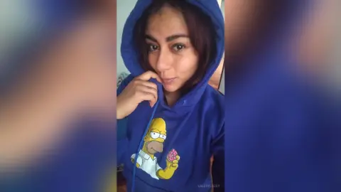 Smiling woman wearing a blue The Simpsons Homer graphic hoodie in a cozy indoor setting.
