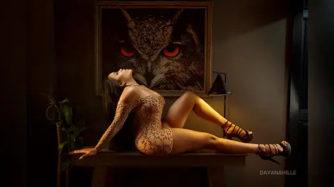 DayanaHille reclines on a wooden table in a tan leopard-print bodysuit, posing against a large owl portrait.