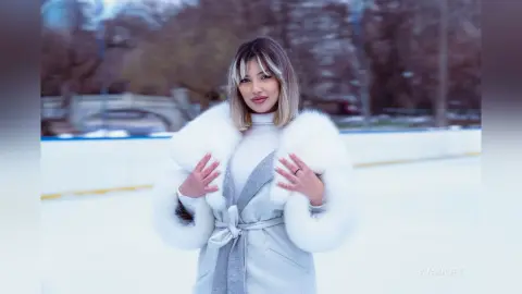 Blonde woman in a white turtleneck and faux fur coat posing on a snowy winter landscape.