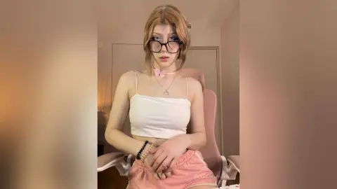 Young woman with glasses in a white crop top and pink shorts sitting in a cozy indoor setting.
