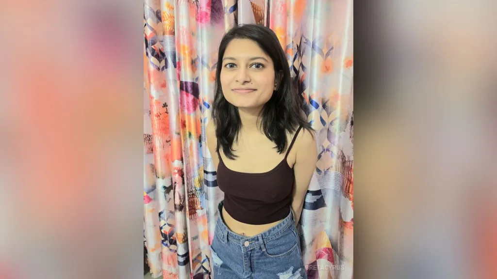 Smiling young woman in a pink ribbed tank top sitting in a bright room with floral curtains.