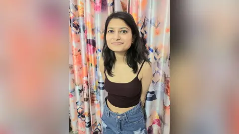 Smiling young woman in a black crop top and denim jeans posing against floral patterned curtains.