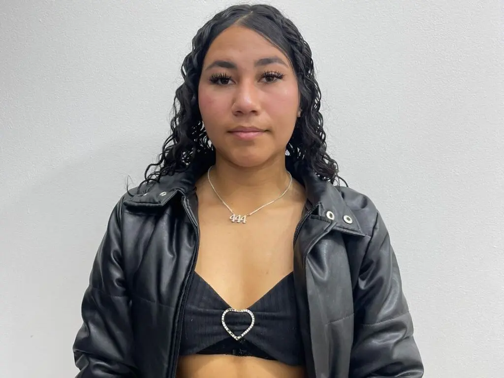 CamilaVelvet poses wearing a black leather jacket and a black tube top with a heart-shaped cutout, showing her long, dark wavy hair against a plain white wall.
